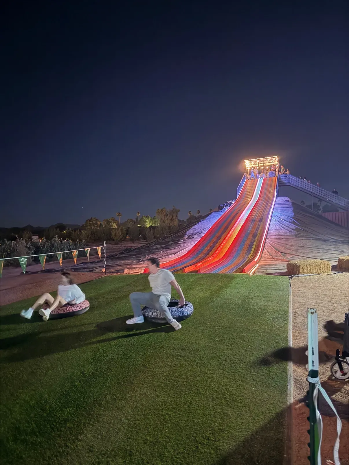 The giant illuminated tube slide at night, riders landing on the turf at the bottom